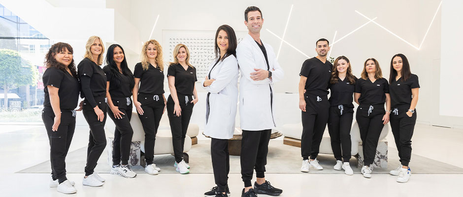 A group of people, likely healthcare professionals, posing in a professional setting with a mix of genders and attire, including scrubs and casual wear.